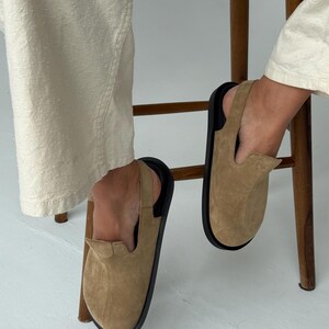 May include: Tan suede slip-on shoes with a black sole and a slingback strap. The shoes are shown with cream-colored pants and a wooden chair.