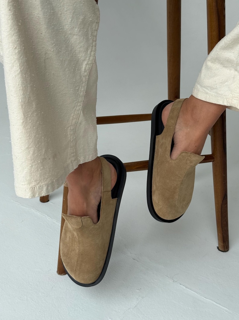 May include: Tan suede slip-on shoes with a black sole and a slingback strap. The shoes are shown with cream-colored pants and a wooden chair.