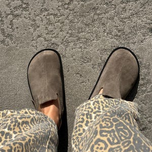 May include: A pair of gray suede slip-on shoes with black soles. The shoes are worn with leopard print pants. The image is taken from a top-down perspective, showing the shoes and the lower legs.