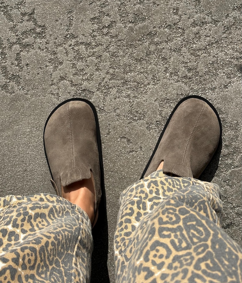 May include: A pair of gray suede slip-on shoes with black soles. The shoes are worn with leopard print pants. The image is taken from a top-down perspective, showing the shoes and the lower legs.
