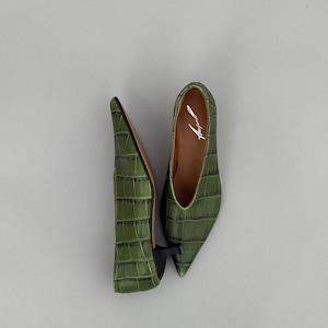 May include: A pair of olive green pointed-toe shoes with a crocodile-embossed texture and a small black kitten heel. The interior of the shoe features a white script logo that reads Azell. The design includes a deep V-cut topline and brown lining.