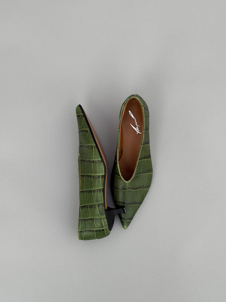 May include: A pair of olive green pointed-toe shoes with a crocodile-embossed texture and a small black kitten heel. The interior of the shoe features a white script logo that reads Azell. The design includes a deep V-cut topline and brown lining.