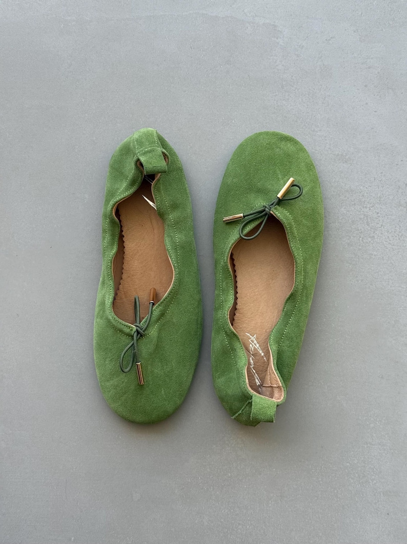 May include: A pair of grass green suede ballet flats featuring a rounded toe, a gathered elasticised topline, and a thin green drawstring cord tied in a bow at the front with metallic gold-toned aglets at the ends of the cord tips.