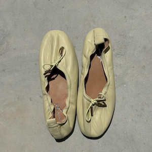 May include: A pair of pale yellow leather ballet flats featuring a soft, gathered construction with a rounded toe. Each shoe includes a delicate drawstring tie at the vamp with metallic gold-toned aglets and a tan interior lining with branding.