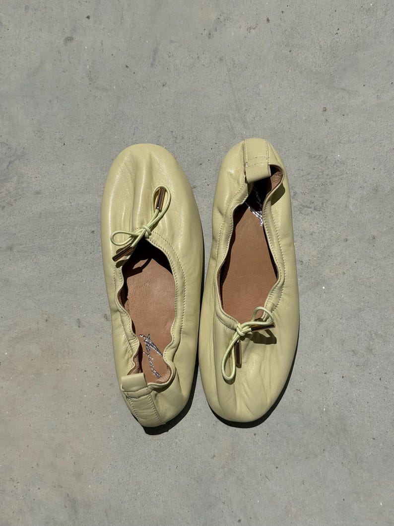 May include: A pair of pale yellow leather ballet flats featuring a soft, gathered construction with a rounded toe. Each shoe includes a delicate drawstring tie at the vamp with metallic gold-toned aglets and a tan interior lining with branding.