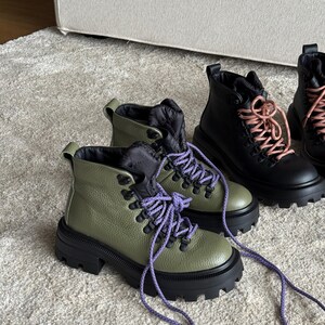 May include: Olive green and black ankle boots with purple laces. The boots have a chunky black sole and a textured leather upper. A pair of black boots with pink laces are also visible.