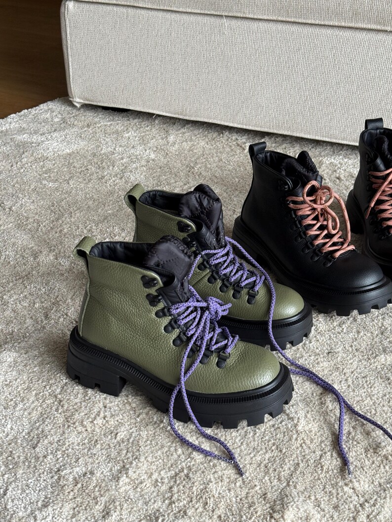 May include: Olive green and black ankle boots with purple laces. The boots have a chunky black sole and a textured leather upper. A pair of black boots with pink laces are also visible.