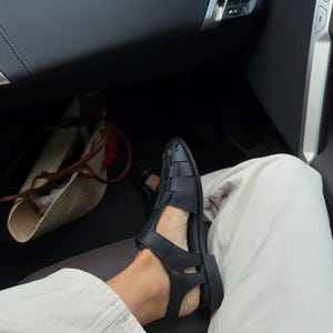 K&ouml;nnte beinhalten: Schwarze Ledersandalen mit einem gewebten Design. Die Sandalen haben eine flache Sohle und werden mit hellen Hosen getragen. Das Bild ist aus einem niedrigen Winkel aufgenommen und zeigt die Sandalen und einen Teil des Beins.