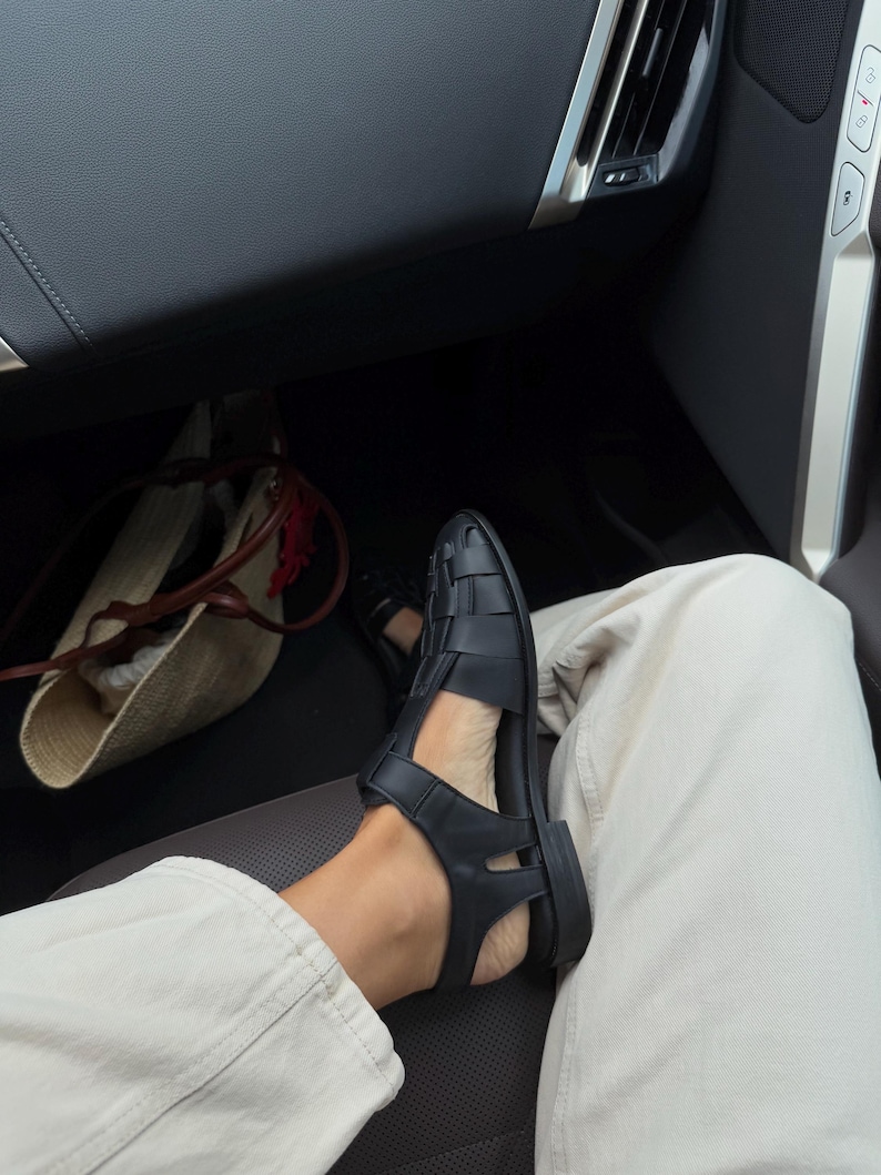 K&ouml;nnte beinhalten: Schwarze Ledersandalen mit einem gewebten Design. Die Sandalen haben eine flache Sohle und werden mit hellen Hosen getragen. Das Bild ist aus einem niedrigen Winkel aufgenommen und zeigt die Sandalen und einen Teil des Beins.
