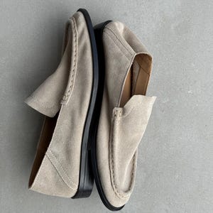 May include: A pair of beige suede loafers with black soles. The shoes have a classic slip-on design with a rounded toe and decorative stitching. The neutral color and simple design make them versatile for various outfits.
