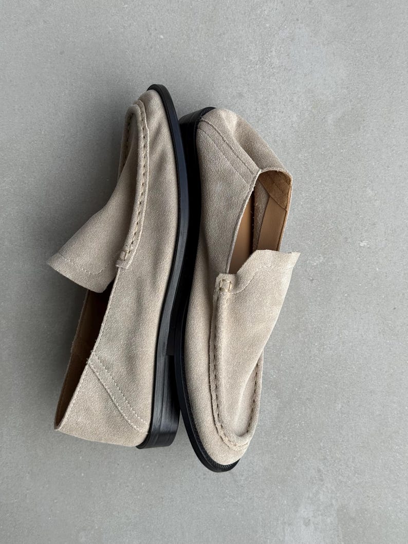 May include: A pair of beige suede loafers with black soles. The shoes have a classic slip-on design with a rounded toe and decorative stitching. The neutral color and simple design make them versatile for various outfits.
