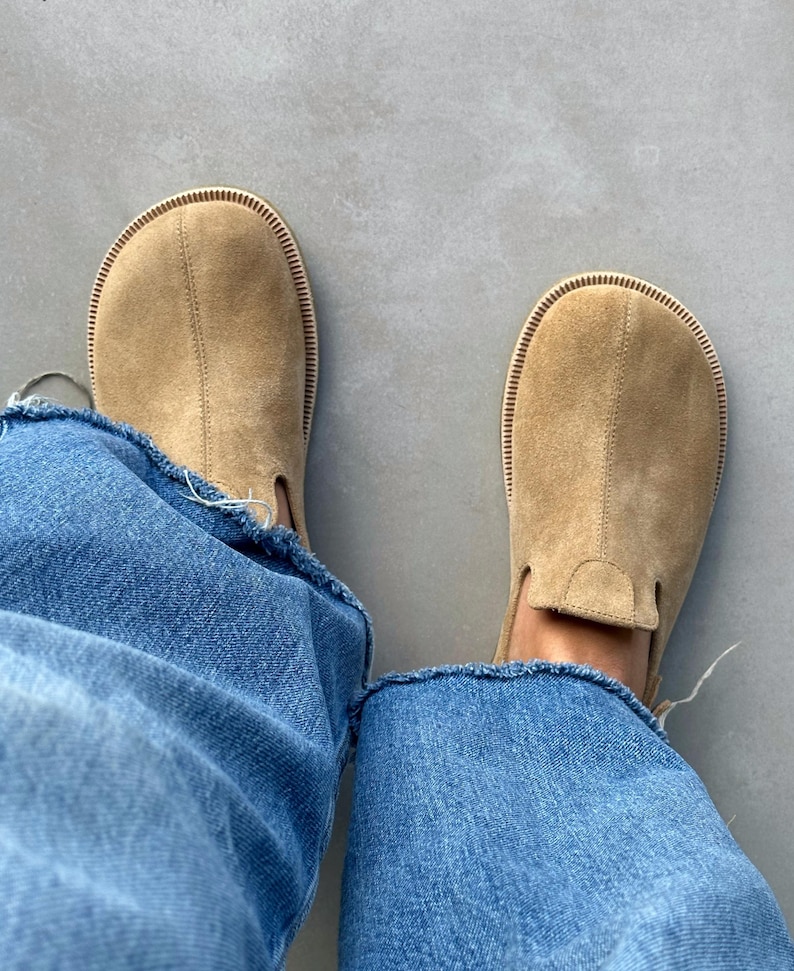 May include: Tan suede slip-on shoes with a rounded toe and stitched edge. The shoes are worn with blue denim jeans with frayed edges. The shoes appear unworn. The shoes are likely size 40-42 EU.