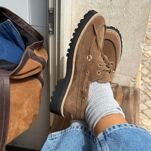 May include: Brown suede boat shoes with a thick black sole. The shoes have a lace-up design and are worn with gray socks and blue jeans. A brown suede tote bag is visible in the background.