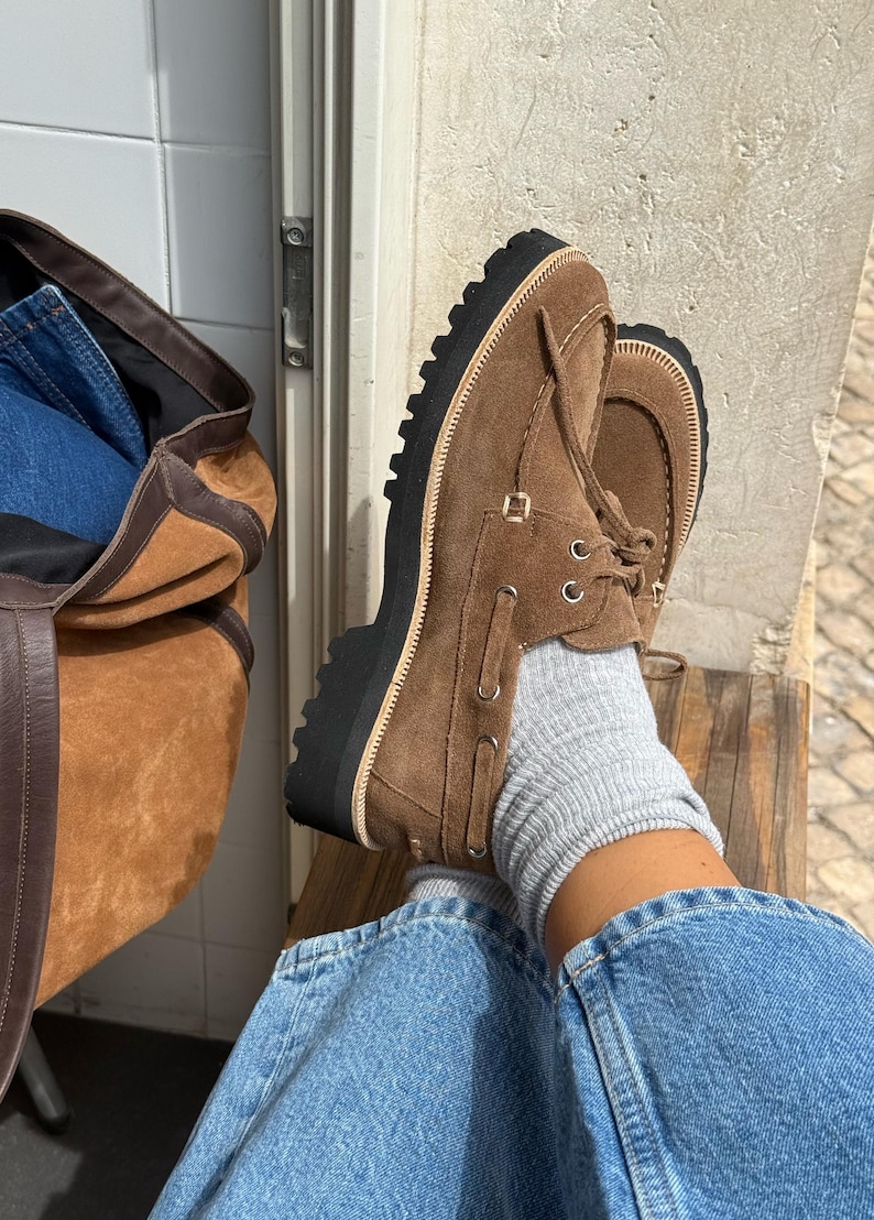 May include: Brown suede boat shoes with a thick black sole. The shoes have a lace-up design and are worn with gray socks and blue jeans. A brown suede tote bag is visible in the background.