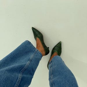 May include: A pair of dark green pointed-toe shoes with a textured crocodile-embossed pattern and a low heel, worn with blue denim jeans. The shoes feature a deep V-cut vamp design and a structured silhouette, providing a sleek look.