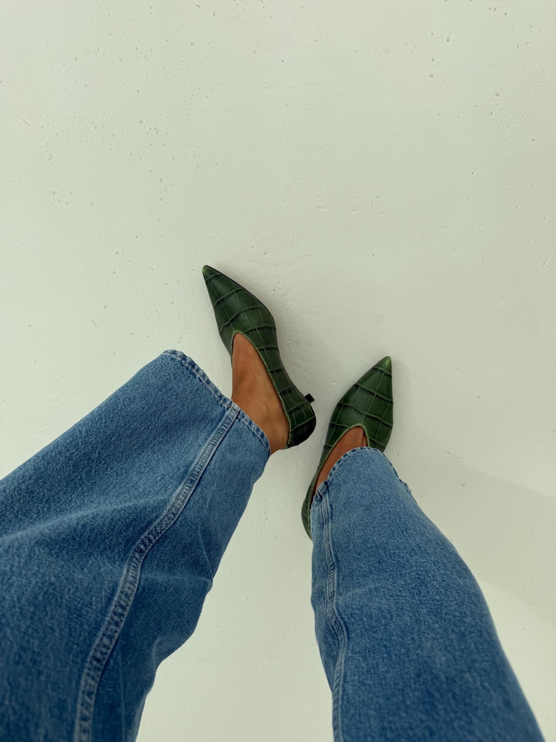 May include: A pair of dark green pointed-toe shoes with a textured crocodile-embossed pattern and a low heel, worn with blue denim jeans. The shoes feature a deep V-cut vamp design and a structured silhouette, providing a sleek look.