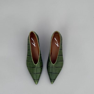 May include: A pair of pointed-toe flats in a deep olive green with a textured crocodile-embossed pattern. The shoes feature a deep V-cut vamp and a brown interior lining with the brand name printed in white script on the heel bed.