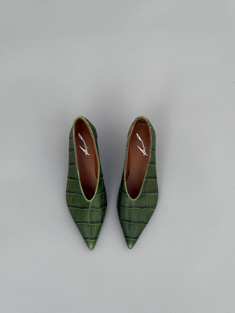 May include: A pair of pointed-toe flats in a deep olive green with a textured crocodile-embossed pattern. The shoes feature a deep V-cut vamp and a brown interior lining with the brand name printed in white script on the heel bed.