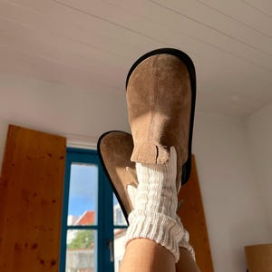 May include: Light brown suede boots with a black sole, paired with cream-coloured knitted leg warmers. The boots have a classic design, and the leg warmers add texture and warmth. The boots are approximately 25 cm tall.
