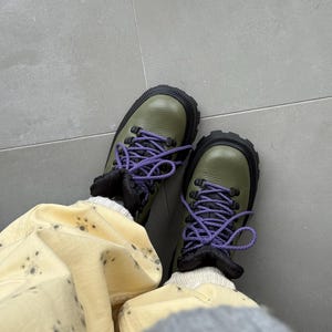 May include: Olive green hiking boots with black soles and purple laces. The boots have a rugged design and are paired with light yellow pants with dark grey spots. The boots appear to be made of leather.