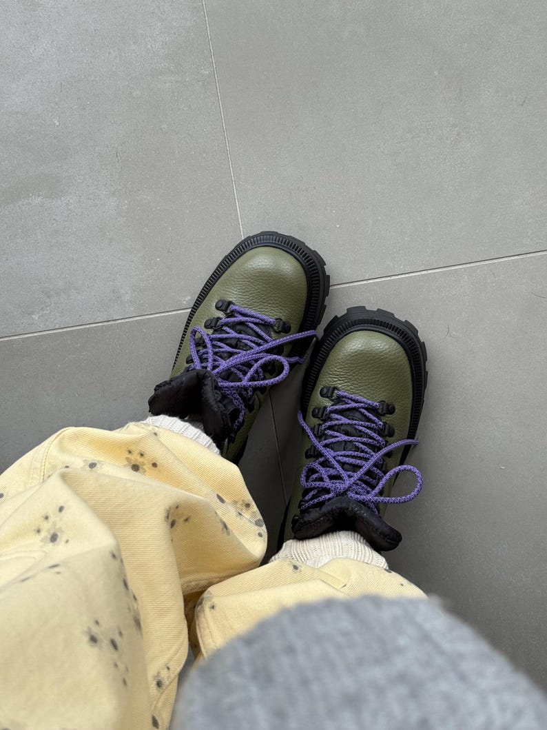 May include: Olive green hiking boots with black soles and purple laces. The boots have a rugged design and are paired with light yellow pants with dark grey spots. The boots appear to be made of leather.