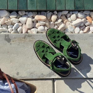 Puede incluir: Un par de sandalias de ante verde con correas trenzadas y cierres de hebilla. Las sandalias tienen suelas negras y se exhiben sobre una superficie de hormig&oacute;n. Una bolsa de paja con tela a rayas azules y blancas est&aacute; en el fondo.
