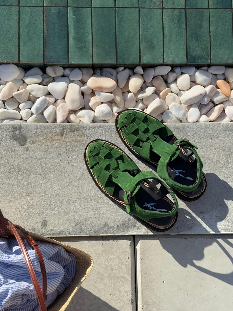 Puede incluir: Un par de sandalias de ante verde con correas trenzadas y cierres de hebilla. Las sandalias tienen suelas negras y se exhiben sobre una superficie de hormig&oacute;n. Una bolsa de paja con tela a rayas azules y blancas est&aacute; en el fondo.