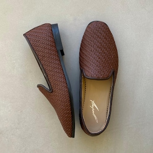 May include: Two brown woven leather loafers. These slip-on shoes feature a rounded toe and a low heel. The interior of one shoe has a printed signature. The shoes are presented on a neutral background.