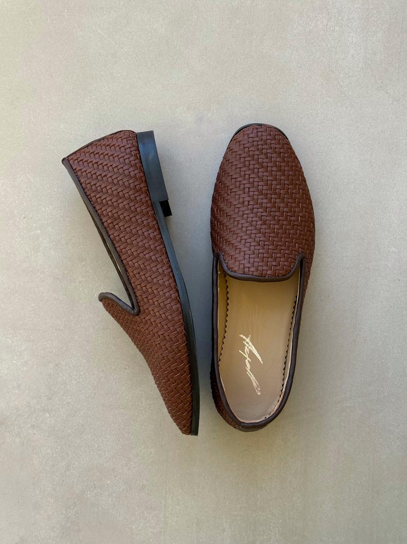 May include: Two brown woven leather loafers. These slip-on shoes feature a rounded toe and a low heel. The interior of one shoe has a printed signature. The shoes are presented on a neutral background.
