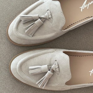 May include: A pair of light beige suede loafers with tassel detailing. The shoes have a rounded toe and a stitched design around the vamp. The soles are a light brown colour, and the interior is a lighter beige. The word "Arper" is written in white on the insole.