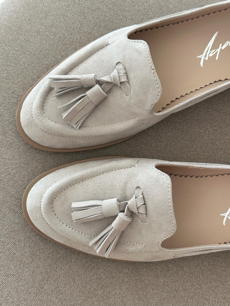 May include: A pair of light beige suede loafers with tassel detailing. The shoes have a rounded toe and a stitched design around the vamp. The soles are a light brown colour, and the interior is a lighter beige. The word "Arper" is written in white on the insole.