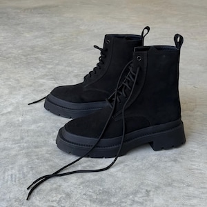 May include: A pair of black suede combat boots with thick, lug soles. The boots have a lace-up design and pull tabs at the top. The boots are displayed on a grey concrete surface.