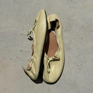May include: A pair of pale yellow leather ballet flats featuring a soft, rounded toe, a gathered elasticised topline, and a decorative bow detail at the front. The interior is lined with a tan material, and the shoes have a thin, dark-coloured sole.