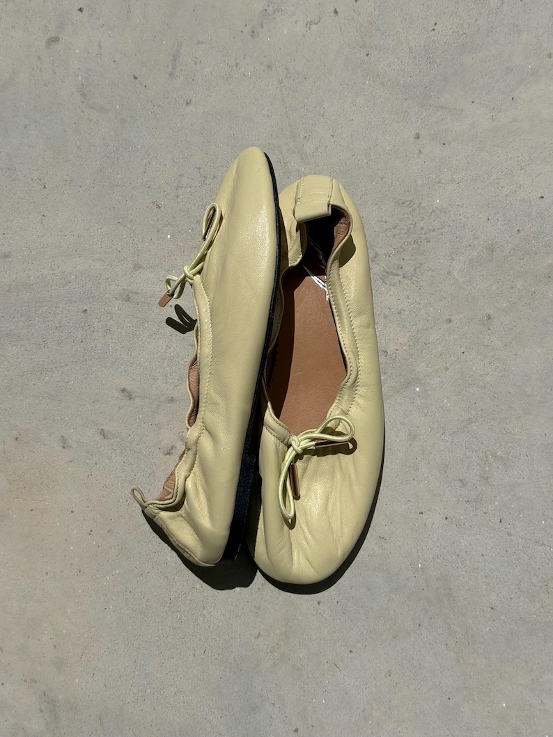May include: A pair of pale yellow leather ballet flats featuring a soft, rounded toe, a gathered elasticised topline, and a decorative bow detail at the front. The interior is lined with a tan material, and the shoes have a thin, dark-coloured sole.