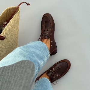 May include: A pair of dark brown leather deck shoes with thin laces and metal eyelets, worn with light blue denim jeans. A woven straw tote bag with brown leather handles is positioned next to the shoes on a neutral grey surface.