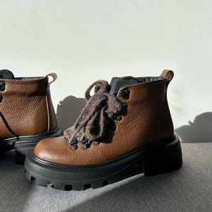 May include: Brown leather ankle boots with black soles and thick, lugged platforms. The boots have dark brown laces and metal eyelets. The boots are designed for outdoor activities and have a rugged, durable appearance.