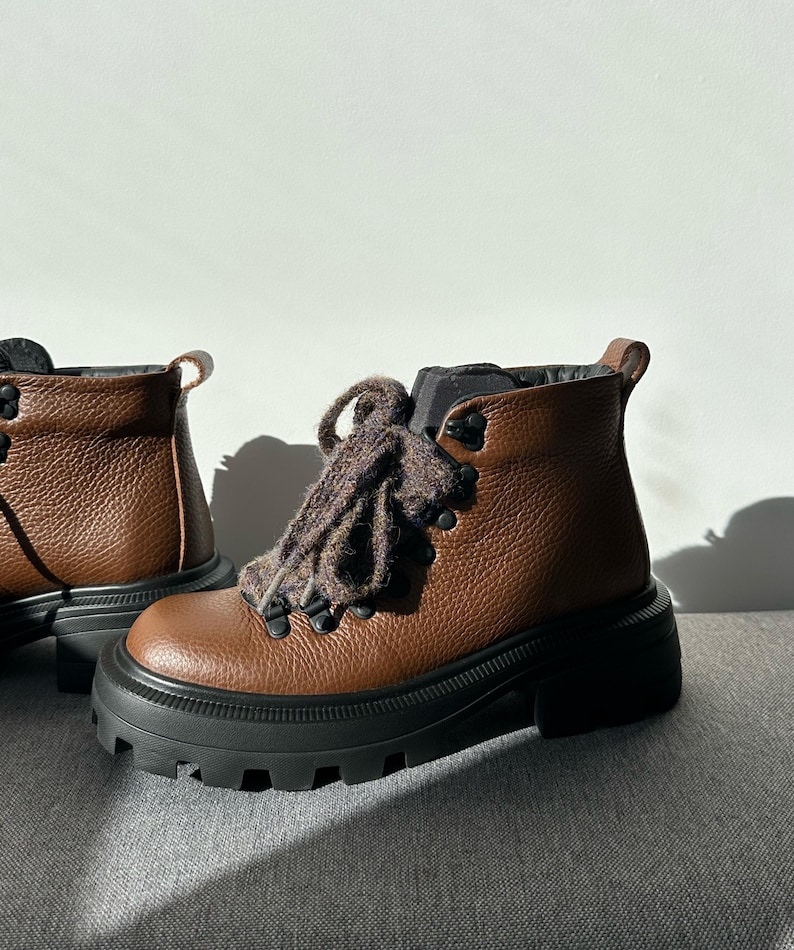 May include: Brown leather ankle boots with black soles and thick, lugged platforms. The boots have dark brown laces and metal eyelets. The boots are designed for outdoor activities and have a rugged, durable appearance.