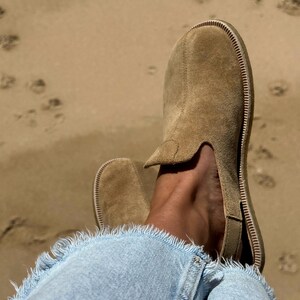 May include: Tan suede slip-on shoes with a stitched sole, worn with light blue denim jeans with frayed edges. The background is a sandy beach with footprints. The shoes are a neutral colour.