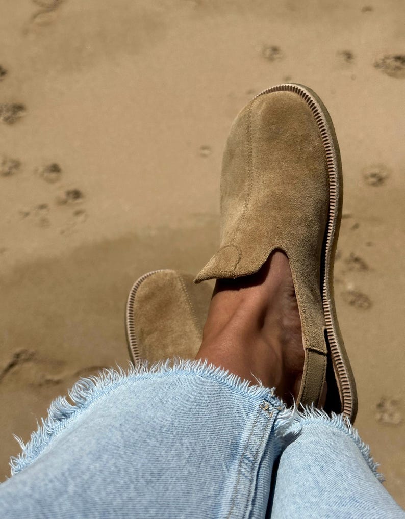 May include: Tan suede slip-on shoes with a stitched sole, worn with light blue denim jeans with frayed edges. The background is a sandy beach with footprints. The shoes are a neutral colour.