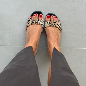 May include: A pair of open-toe slide sandals featuring a textured leopard print pattern on the wide front strap. The sandals have a black footbed and a low profile design. The footwear is paired with dark gray trousers and a silver toe ring.