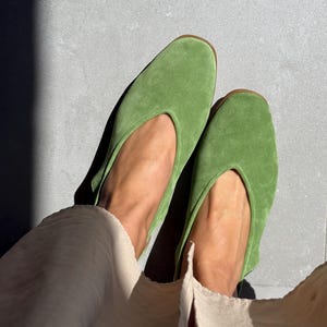 Puede incluir: Un par de bailarinas de ante verde con punta redondeada y escote bajo. Los zapatos tienen una suela marrón claro. Las bailarinas son de un tono verde vibrante, y el estilo general es minimalista y elegante.