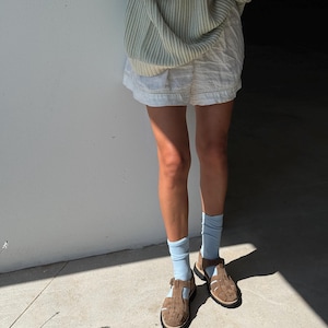 Puede incluir: Una persona con un su&eacute;ter a rayas verde claro, pantalones cortos blancos, calcetines azul claro y sandalias marrones. Las sandalias tienen un dise&ntilde;o tejido y cierre de hebilla. El atuendo es casual y veraniego.