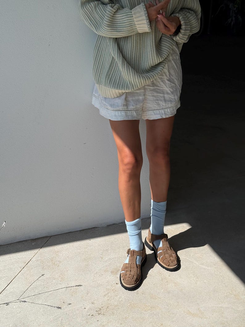 Puede incluir: Una persona con un su&eacute;ter a rayas verde claro, pantalones cortos blancos, calcetines azul claro y sandalias marrones. Las sandalias tienen un dise&ntilde;o tejido y cierre de hebilla. El atuendo es casual y veraniego.