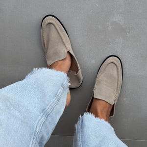 May include: A pair of beige suede loafers with a rounded toe and a black sole. The shoes are worn with light blue denim jeans with frayed edges. The image is taken from a top-down perspective.