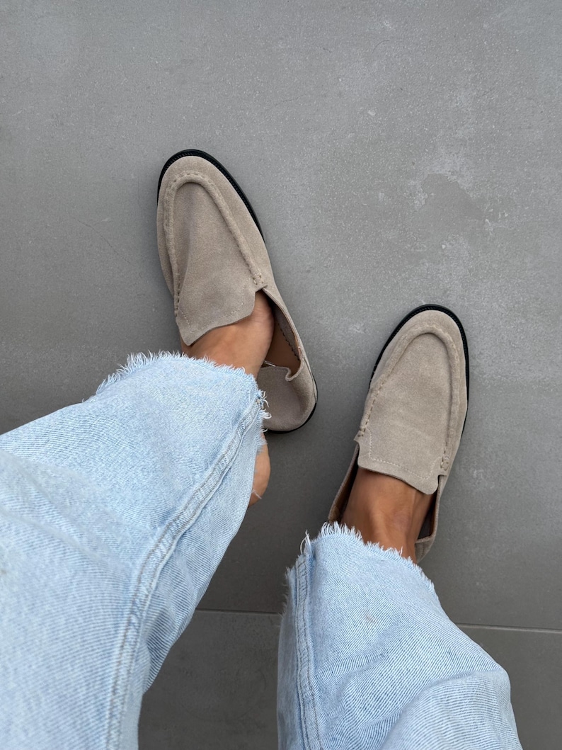 May include: A pair of beige suede loafers with a rounded toe and a black sole. The shoes are worn with light blue denim jeans with frayed edges. The image is taken from a top-down perspective.