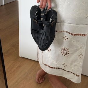 K&ouml;nnte beinhalten: Schwarze Sandalen mit geflochtenem Design und Schnallenverschl&uuml;ssen. Die Sandalen werden von einer Hand gehalten. Sie haben einen Riemen um den Kn&ouml;chel und einen kleinen Absatz. Hergestellt aus synthetischem Material.