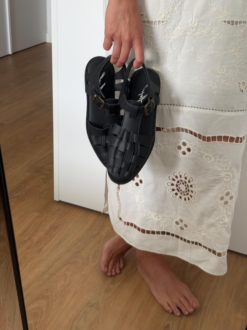 K&ouml;nnte beinhalten: Schwarze Sandalen mit geflochtenem Design und Schnallenverschl&uuml;ssen. Die Sandalen werden von einer Hand gehalten. Sie haben einen Riemen um den Kn&ouml;chel und einen kleinen Absatz. Hergestellt aus synthetischem Material.