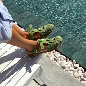 Puede incluir: Sandalias verdes de cuero tejido con correas en el tobillo. Las sandalias tienen suela plana y un dise&ntilde;o tejido en la parte superior. Las sandalias son de color verde claro. El fondo incluye una piscina y una tumbona blanca.