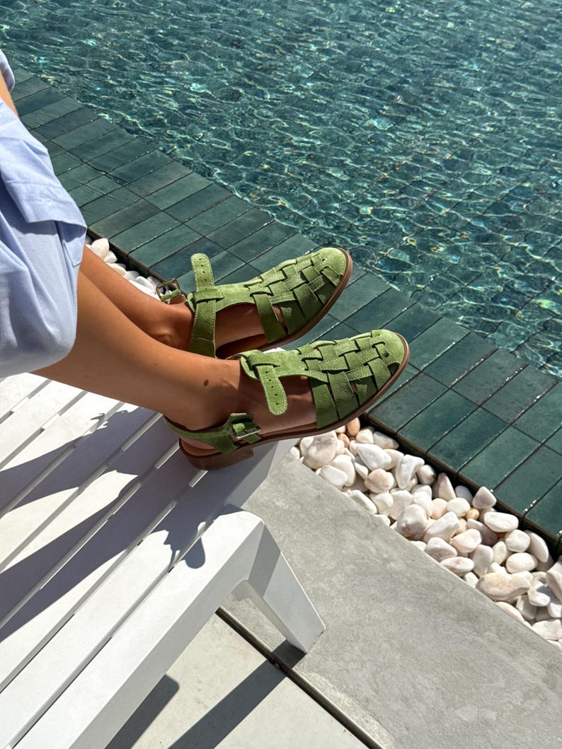 Puede incluir: Sandalias verdes de cuero tejido con correas en el tobillo. Las sandalias tienen suela plana y un dise&ntilde;o tejido en la parte superior. Las sandalias son de color verde claro. El fondo incluye una piscina y una tumbona blanca.
