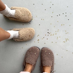 Pode incluir: Vista aérea de dois pares de chinelos estilo shearling, bege e castanho, com enfeites de estrela. Um par é usado com meias brancas. Confetes dourados em forma de estrela espalhados na superfície cinza. Calçado confortável.